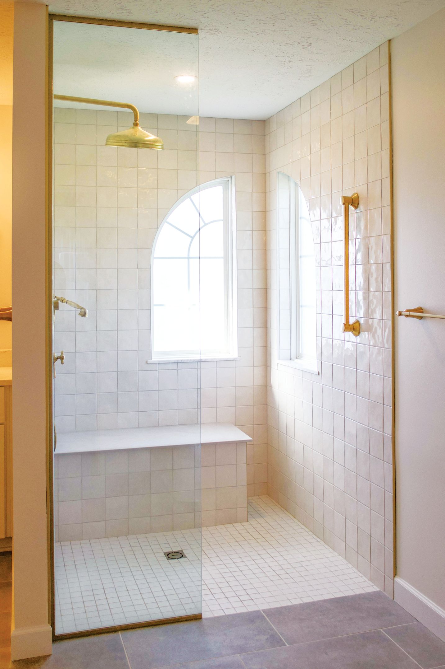 A walk-in shower with white textured tile walls, a glass door, a built-in bench, and gold-toned fixtures.