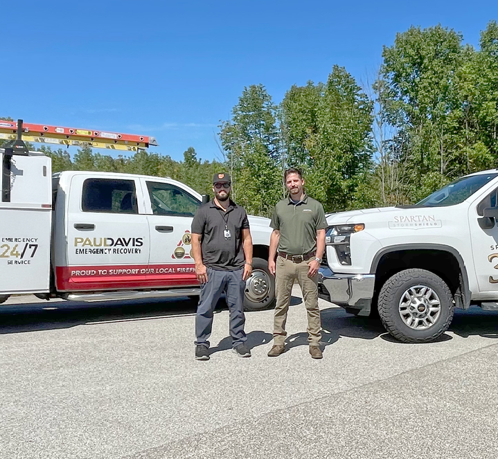 Two workers stand in a gravel lot between two Paul Davis restoration service trucks on a sunny day.