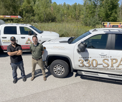 Two people stand outside in front of two white service trucks parked near trees on a sunny day.