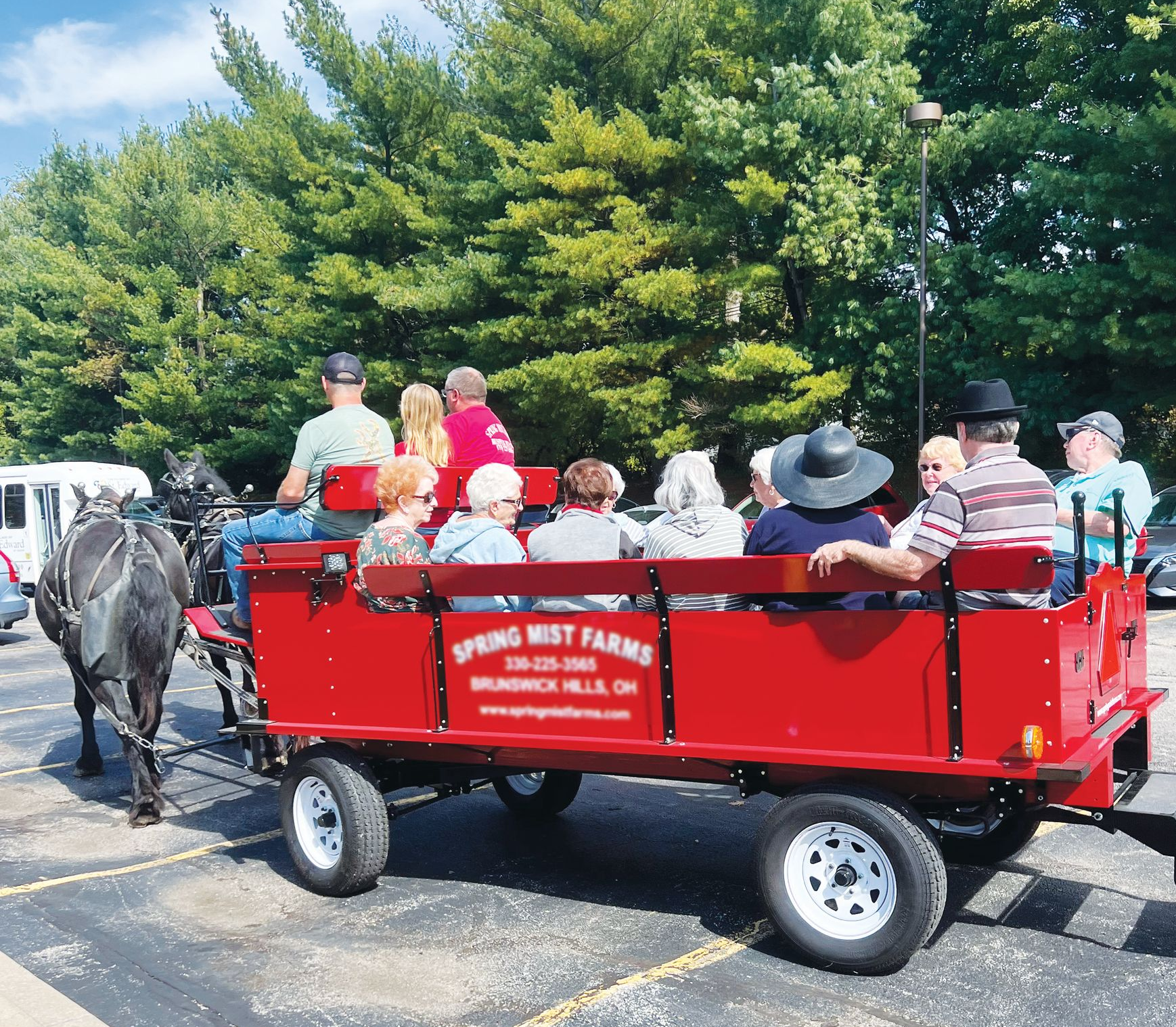 A horse-drawn red carriage carrying several people through a parking lot with a line of trees in the background.