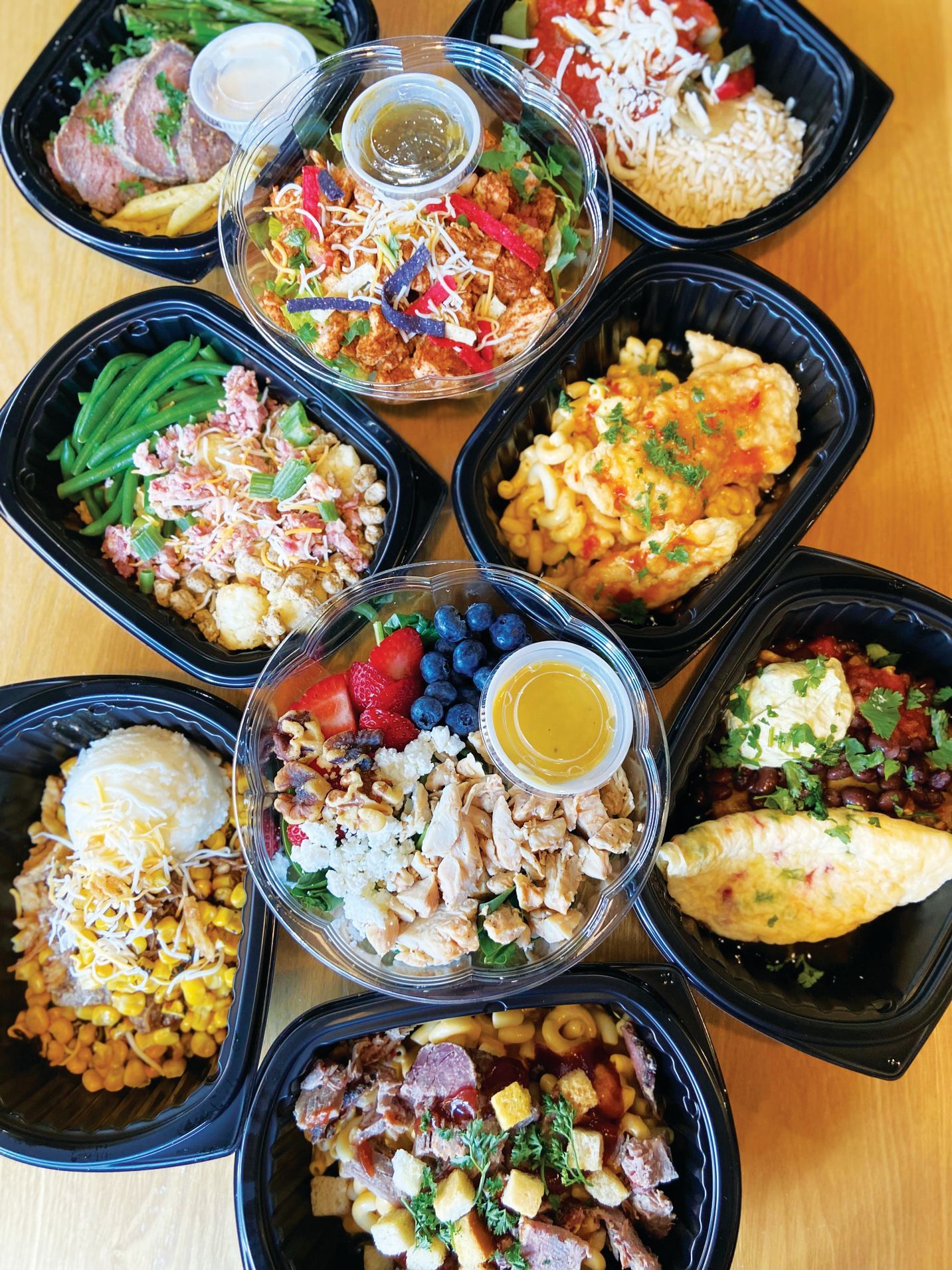 A high-angle view of eight assorted prepared meals in black containers arranged on a light-colored wooden table.