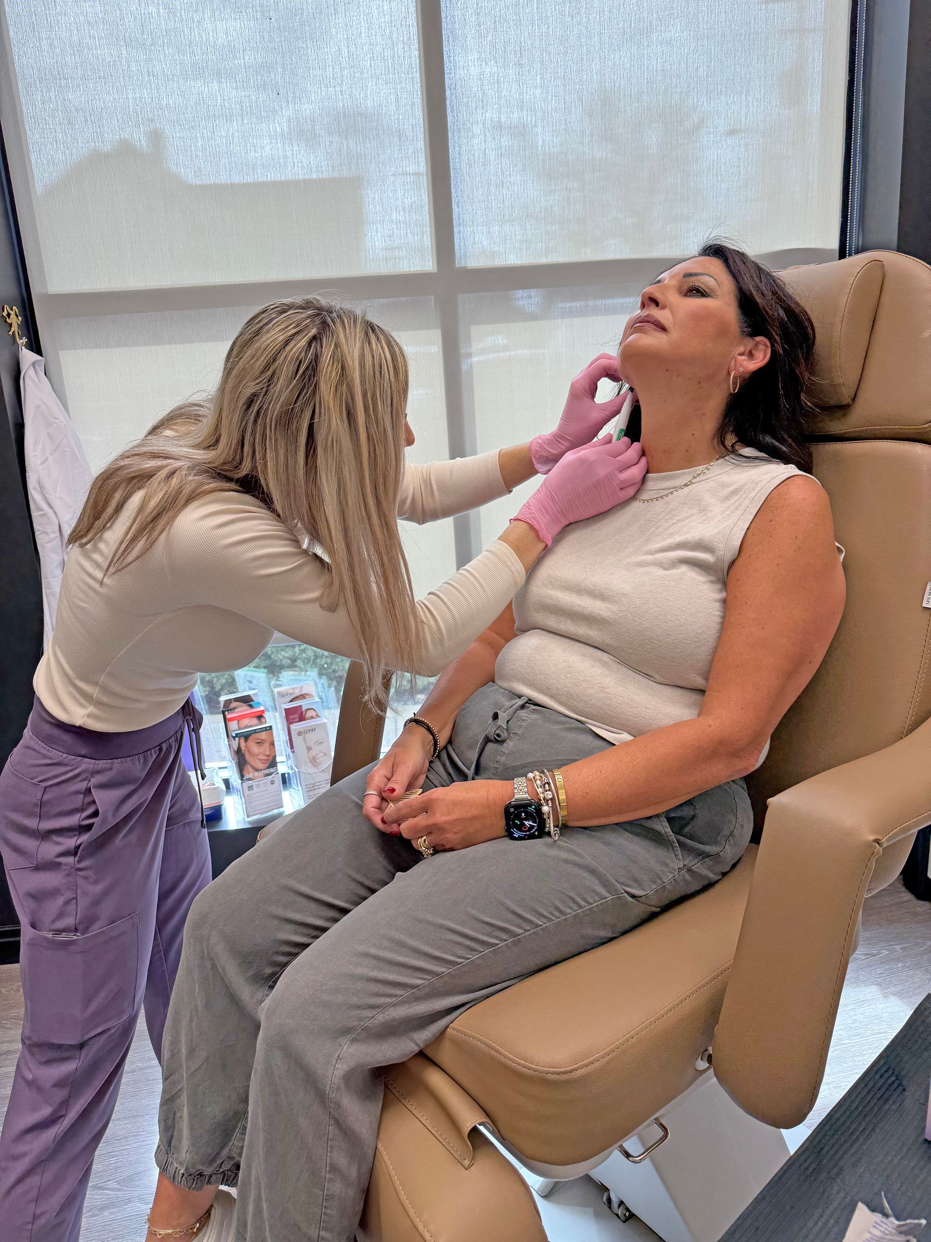 A practitioner in pink gloves administers a cosmetic injection to a person's neck while they sit in a medical chair.