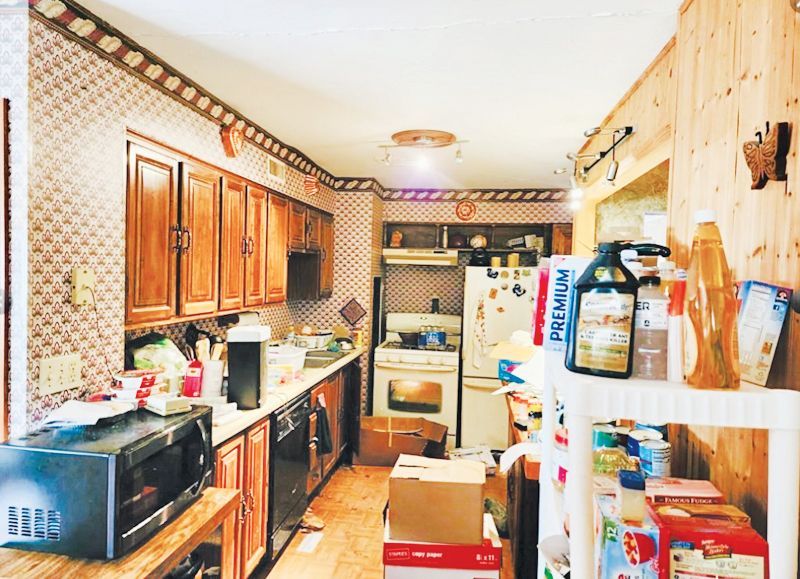 A cluttered kitchen with wooden cabinets, patterned wallpaper, a microwave, white appliances, and boxes on the floor.