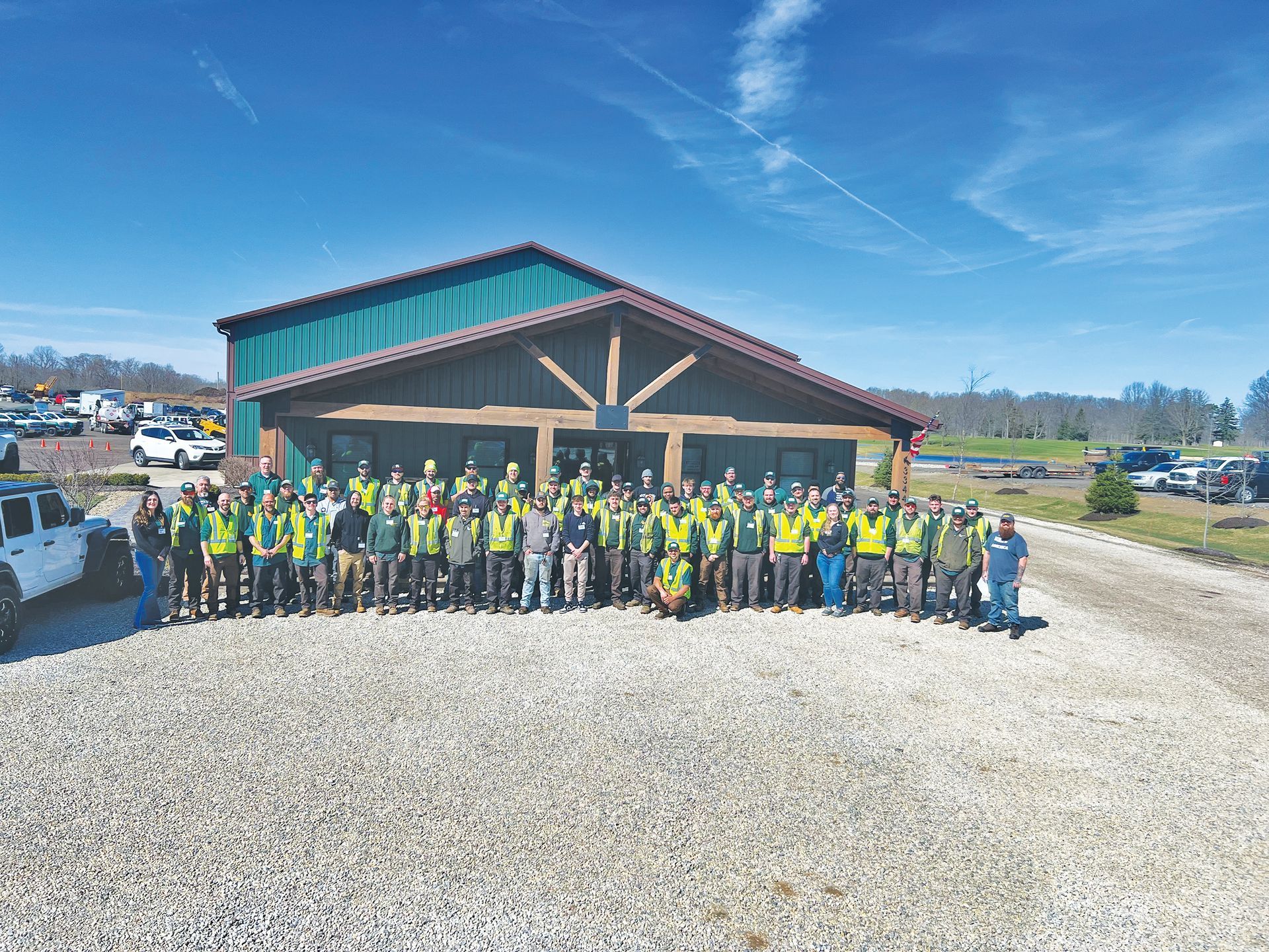 A large group of people in high-visibility vests posing for a photo in front of a building on a gravel lot under a blue sky.