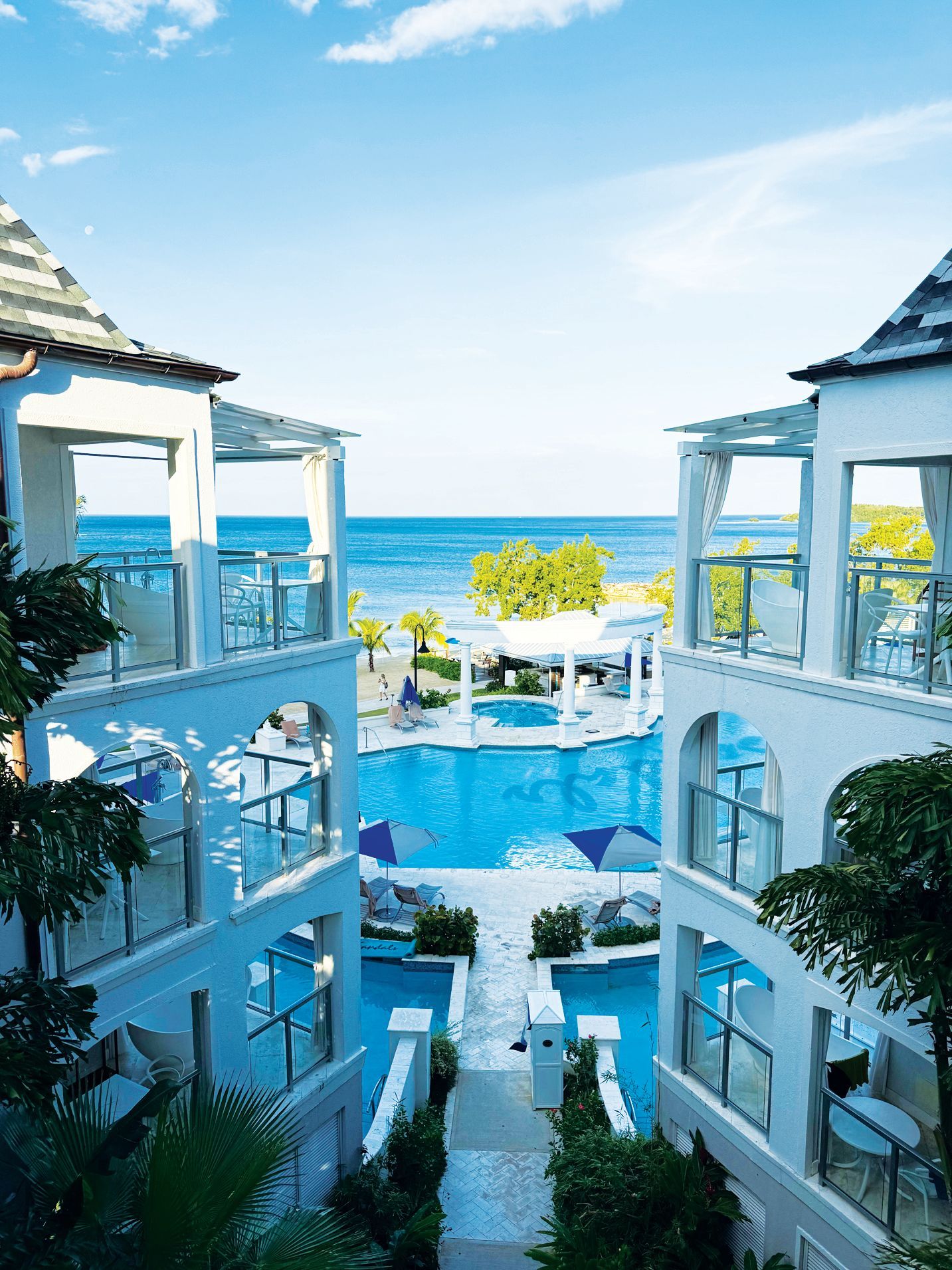 White resort balconies overlook a blue swimming pool with ocean views in the background under a bright, sunny sky.
