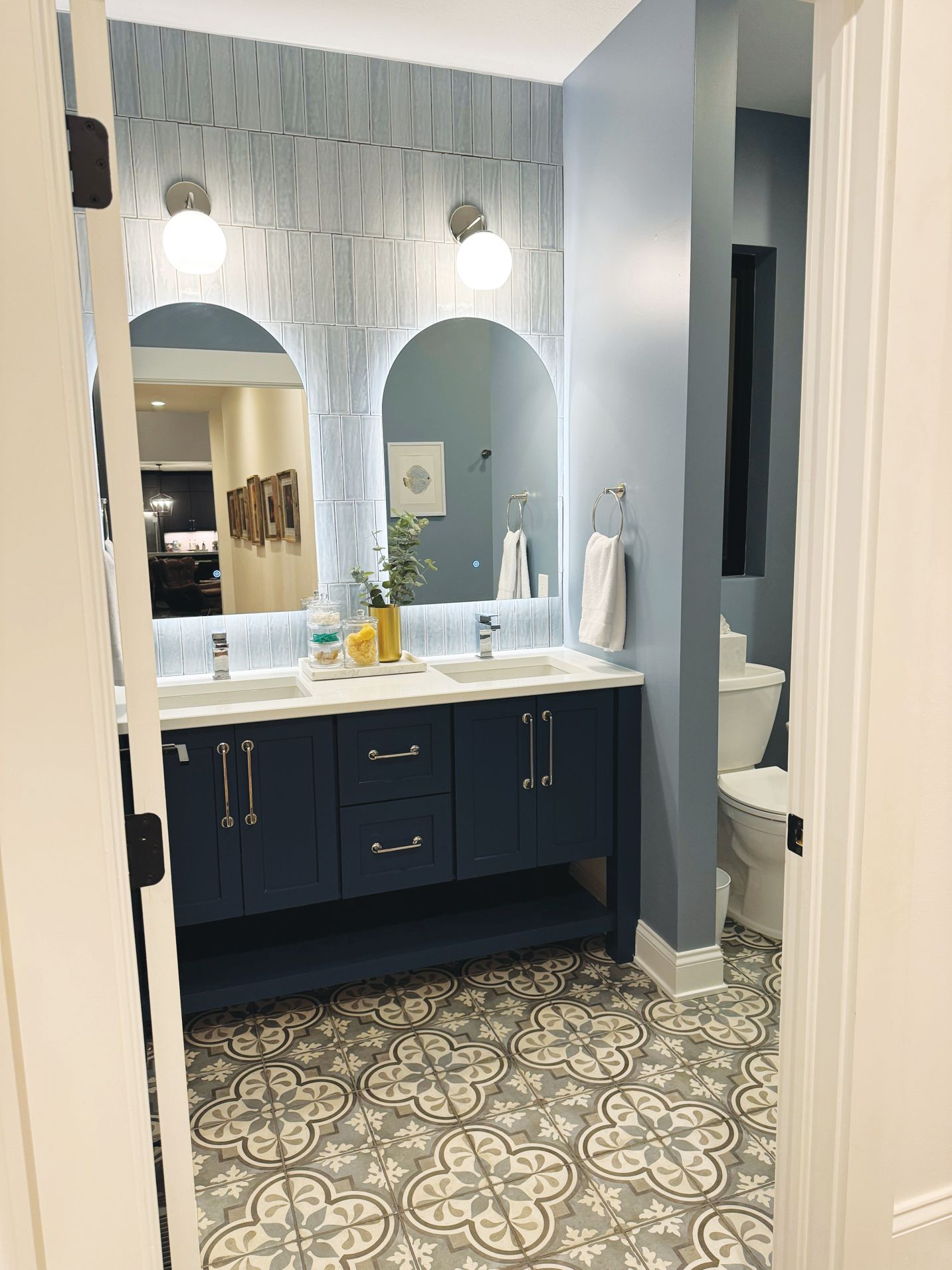 A bathroom with a navy blue vanity, patterned tile floor, two arched mirrors, and light blue walls.