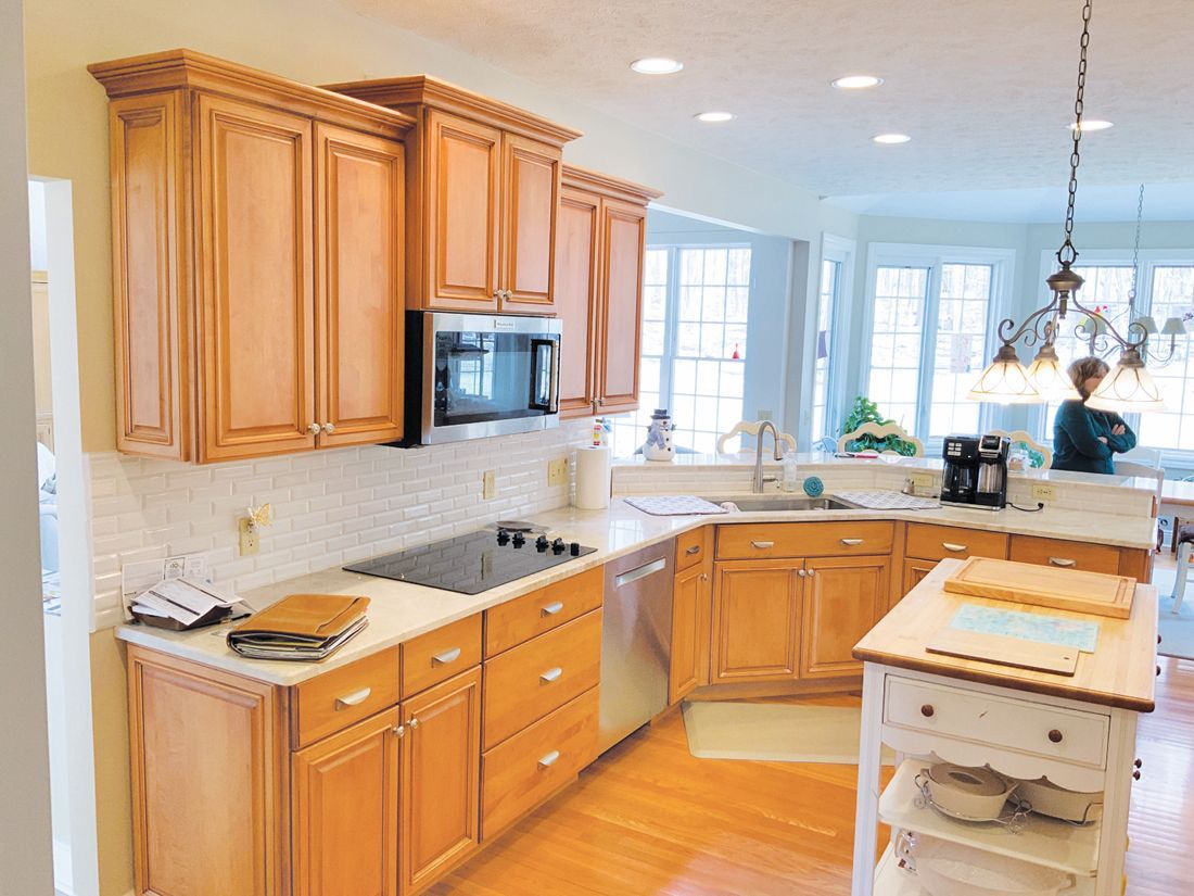 A bright kitchen features light wood cabinets, white countertops, a central island, and a person standing in the background.