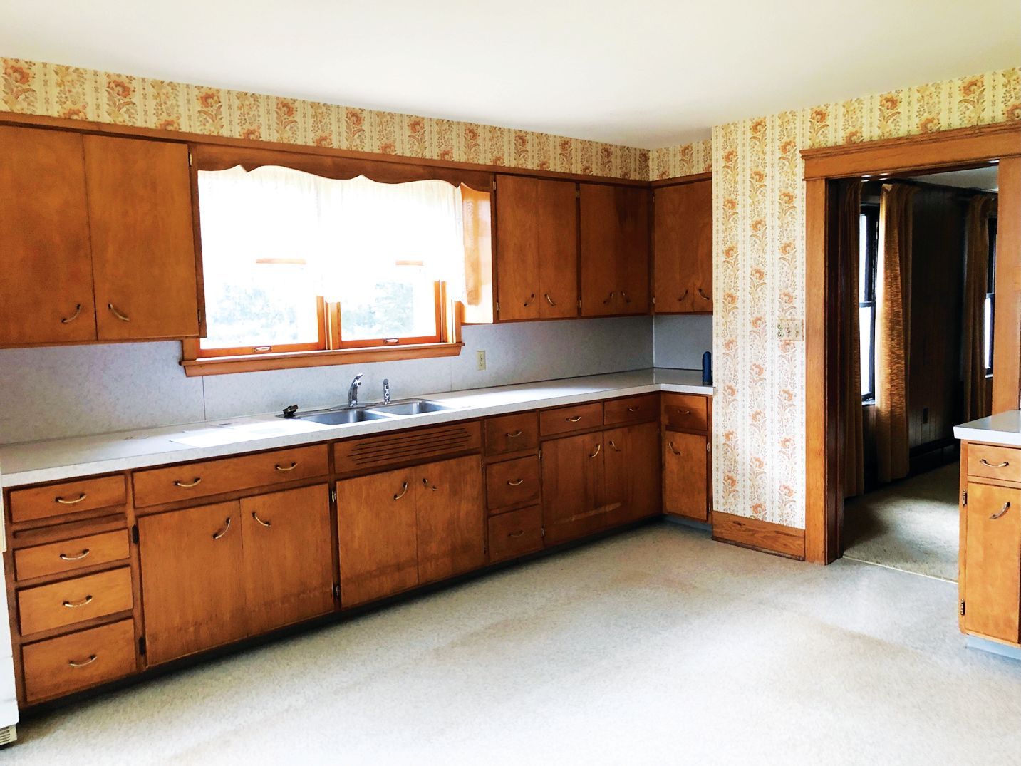 A vintage kitchen with wood cabinets, light-colored countertops, and floral wallpaper above the cabinets and doorway.