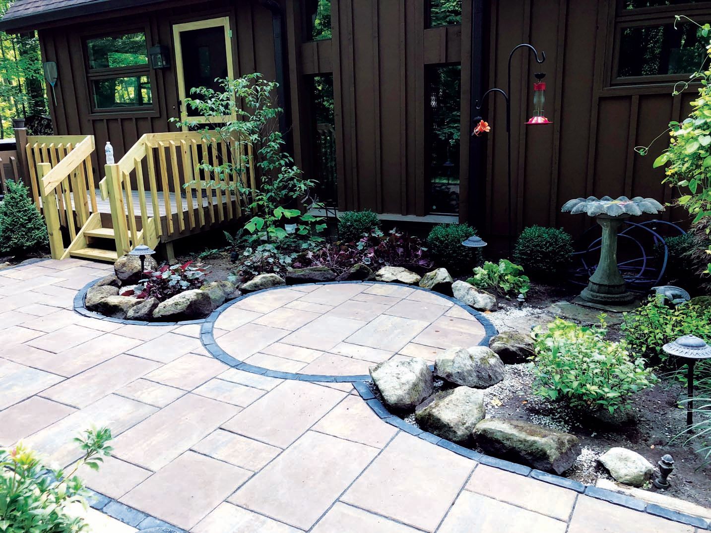 A stone patio with a circular inlay feature leads to a wooden deck beside a brown house with landscaping and a birdbath.