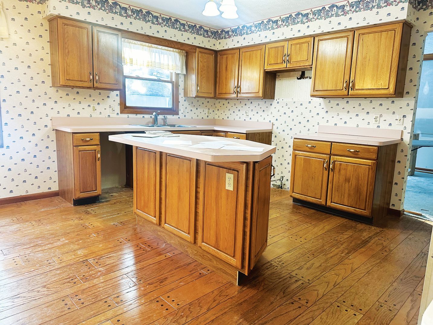 A kitchen with honey-oak cabinets, a central island, light-colored countertops, and patterned wallpaper over wood floors.