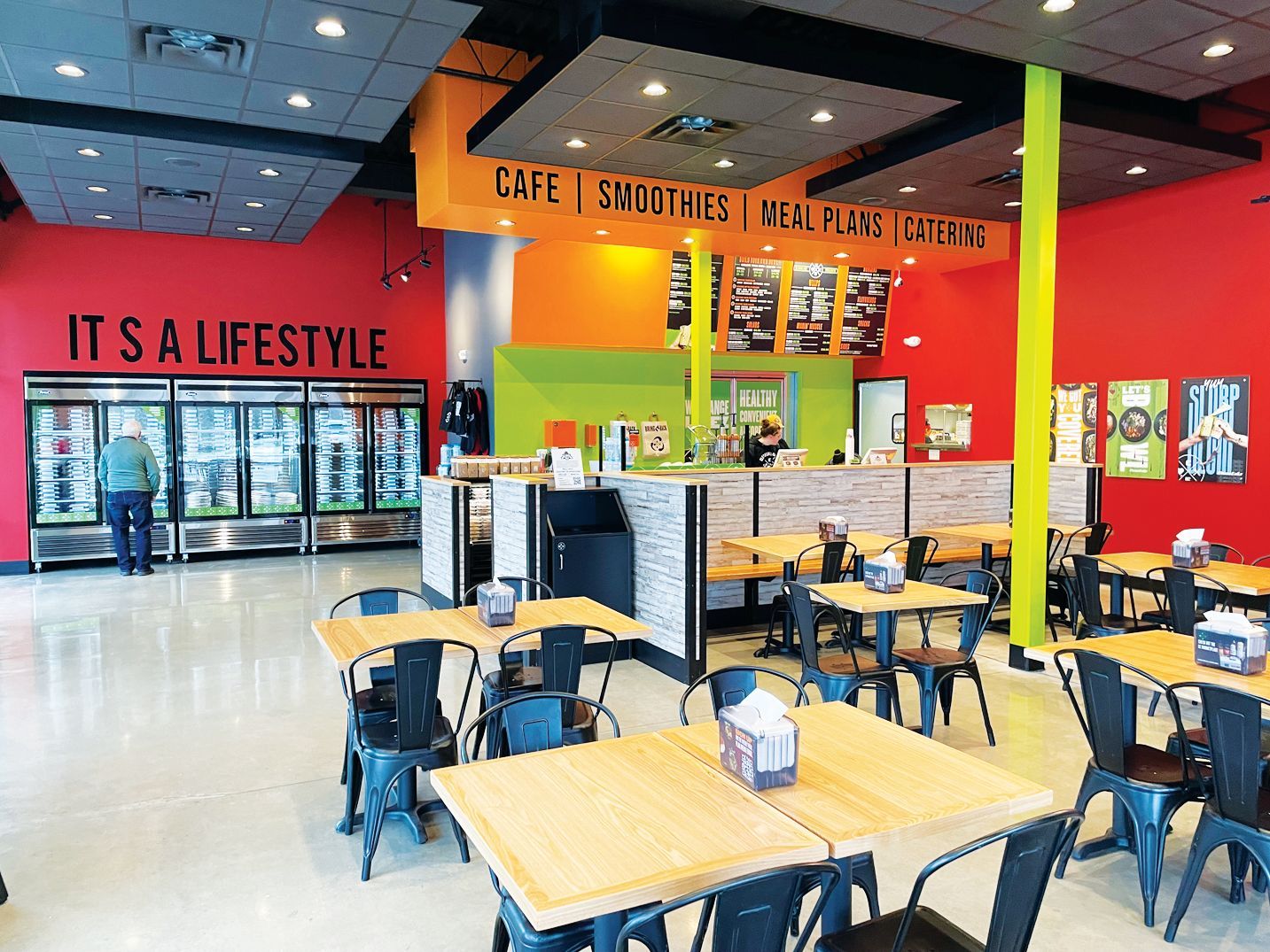 An interior view of a vibrant cafe with red and orange walls, light wood tables, black chairs, and a serving counter.