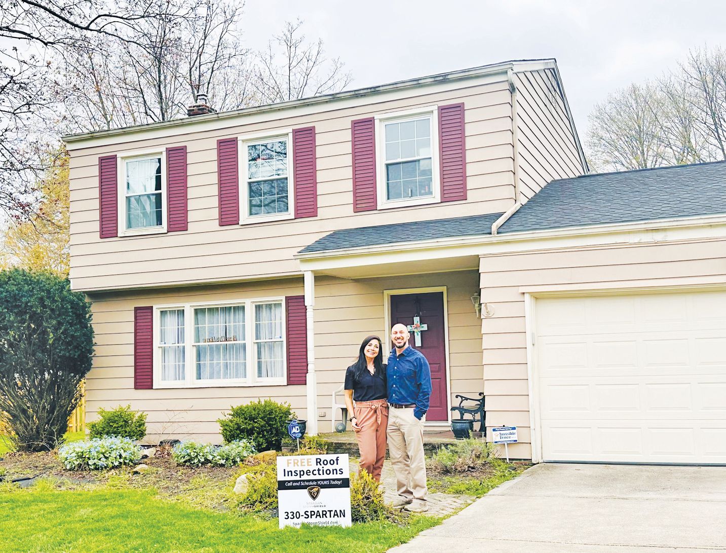 A couple stands smiling in front of their suburban home, with a