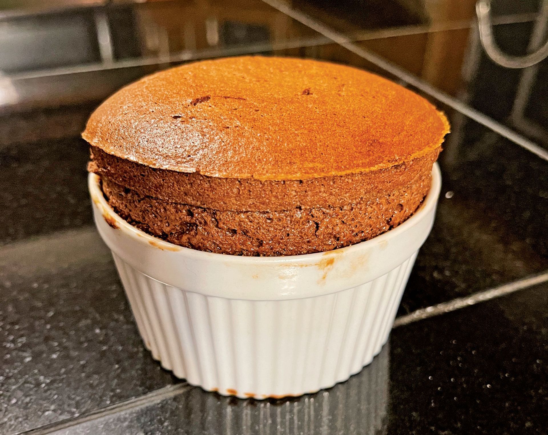 A chocolate soufflé rises perfectly in a small, white, ribbed ceramic ramekin, set on a dark, reflective counter.