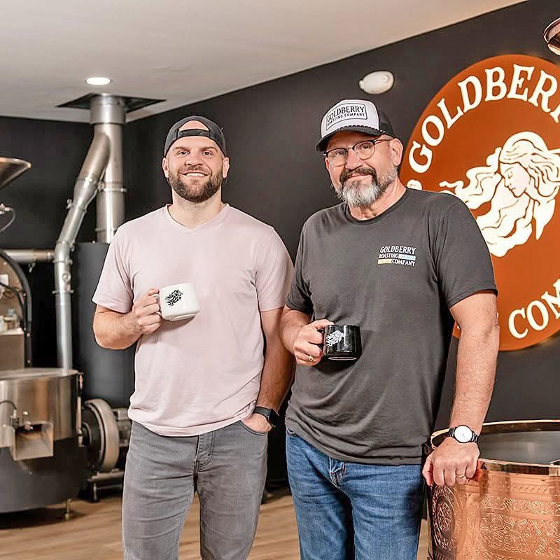 Two men holding coffee mugs stand in front of a Goldberry Coffee Company sign and roasting equipment.