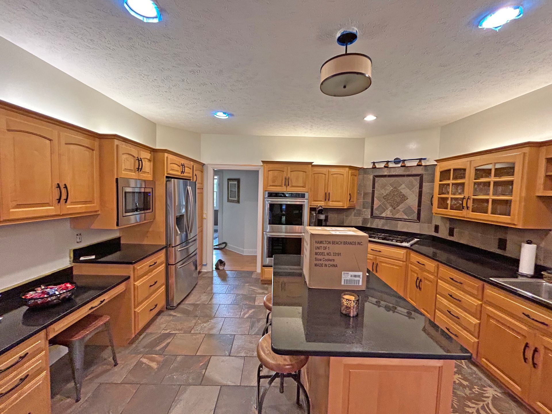 A kitchen with light wood cabinets, dark granite countertops, stainless steel appliances, and large stone floor tiles.