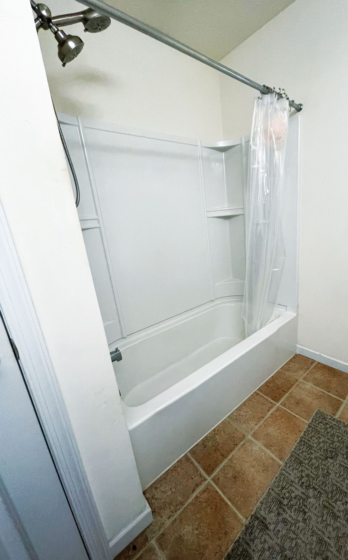 A white bathtub and shower surround with a metal rod and clear plastic curtain, set in a room with tan tile flooring.