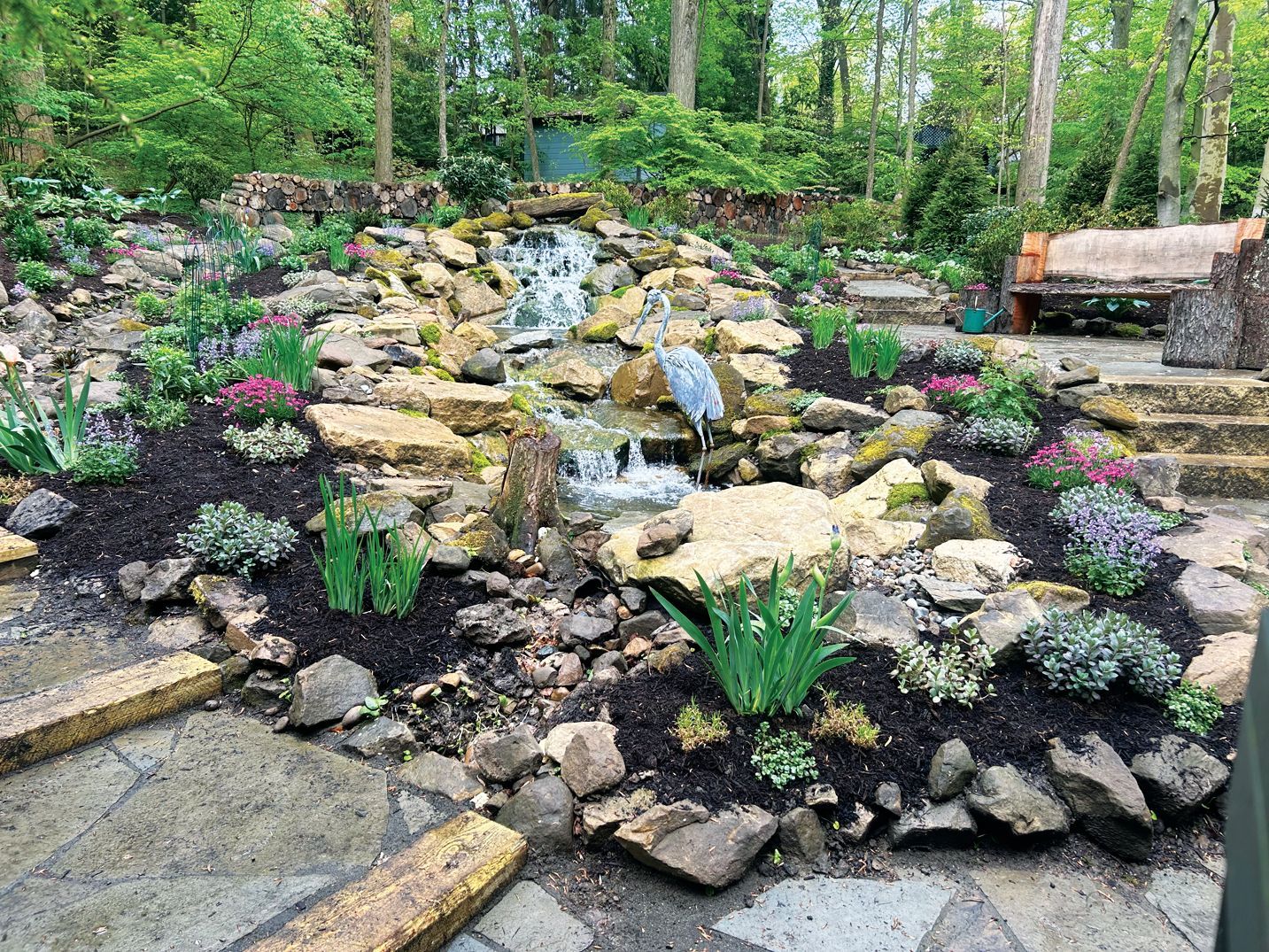 A stone waterfall cascades into a small pond in a lush garden, featuring decorative plants and a wooden bench nearby.