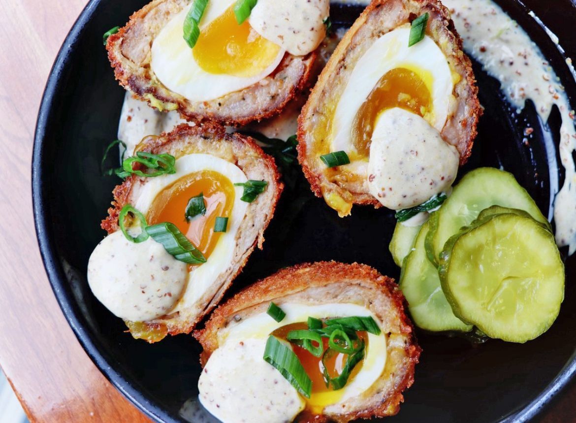 Four halved Scotch eggs with soft yolks, garnished with green onions and sauce, served with pickles in a black bowl.