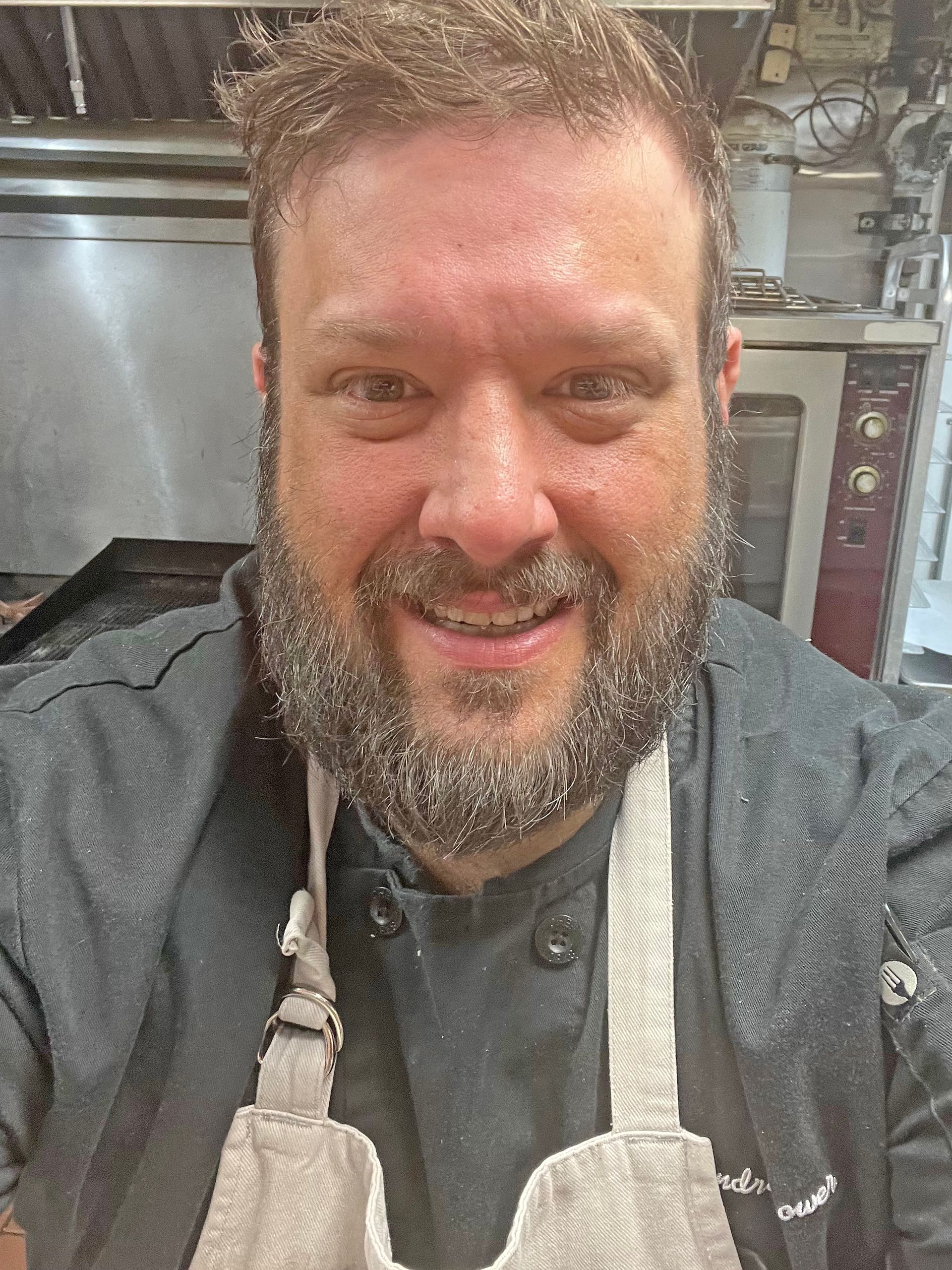A smiling person with a beard, wearing a black chef’s coat and a light-colored apron, stands in a kitchen.