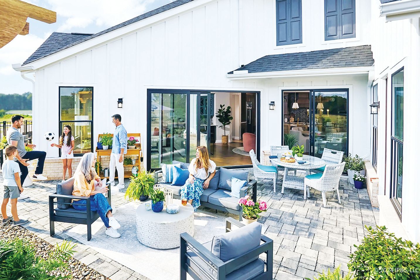 A group of people gathers on a sunny stone patio outside a modern white home, relaxing on outdoor furniture near an open door.