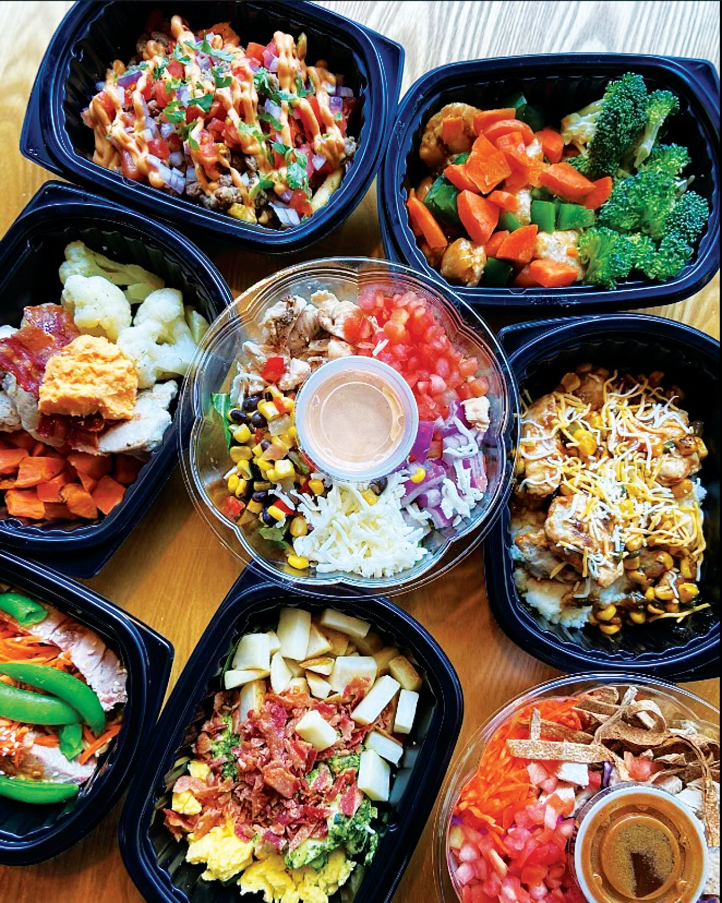 An overhead view of several colorful, healthy meal prep containers with vegetables, proteins, and grains on a table.