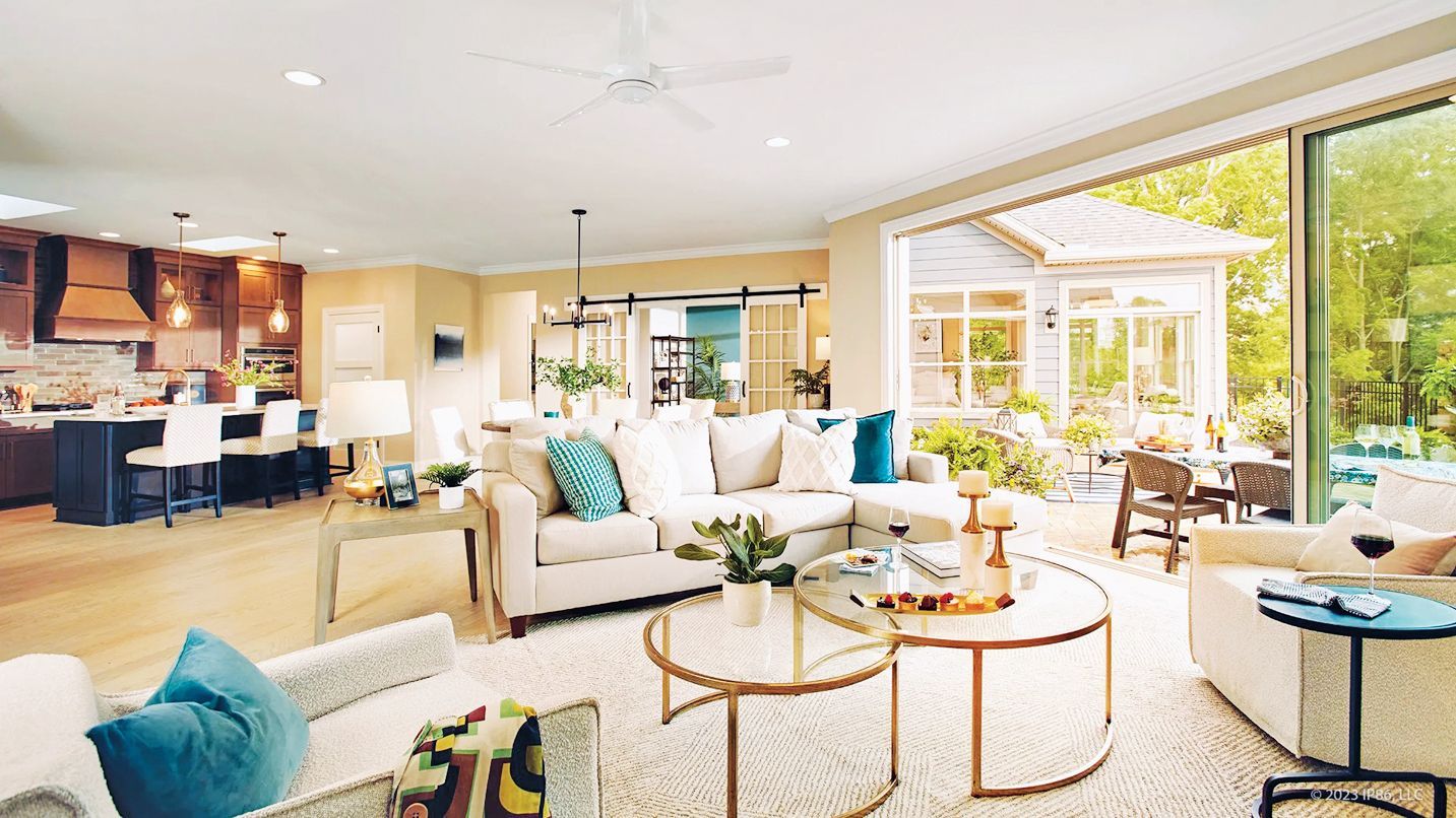 A bright, open-plan living room with white sofas, a central coffee table, and an adjacent kitchen leading to a patio.