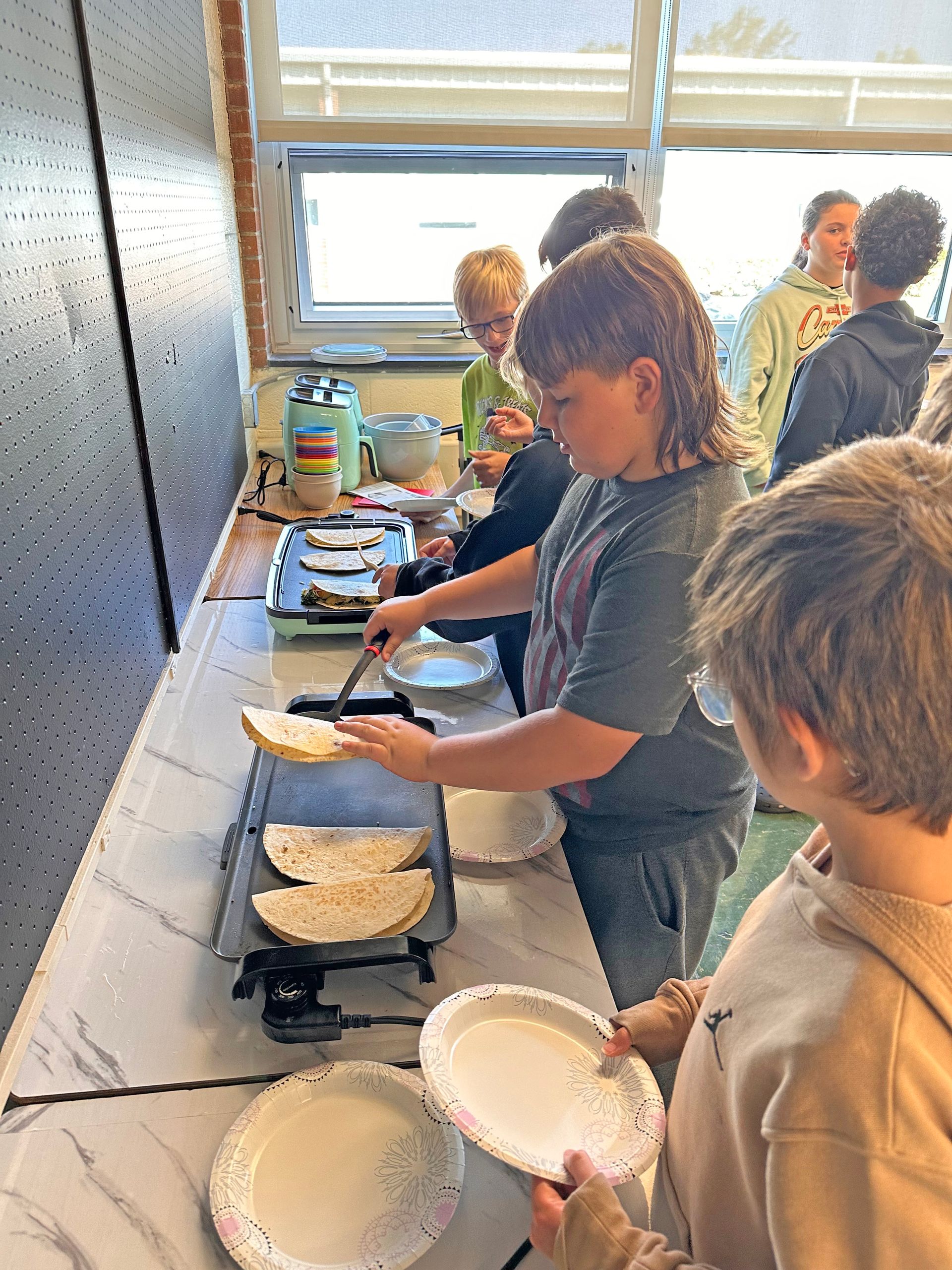 Students cook tortillas on an electric griddle in a classroom setting, preparing food on paper plates.
