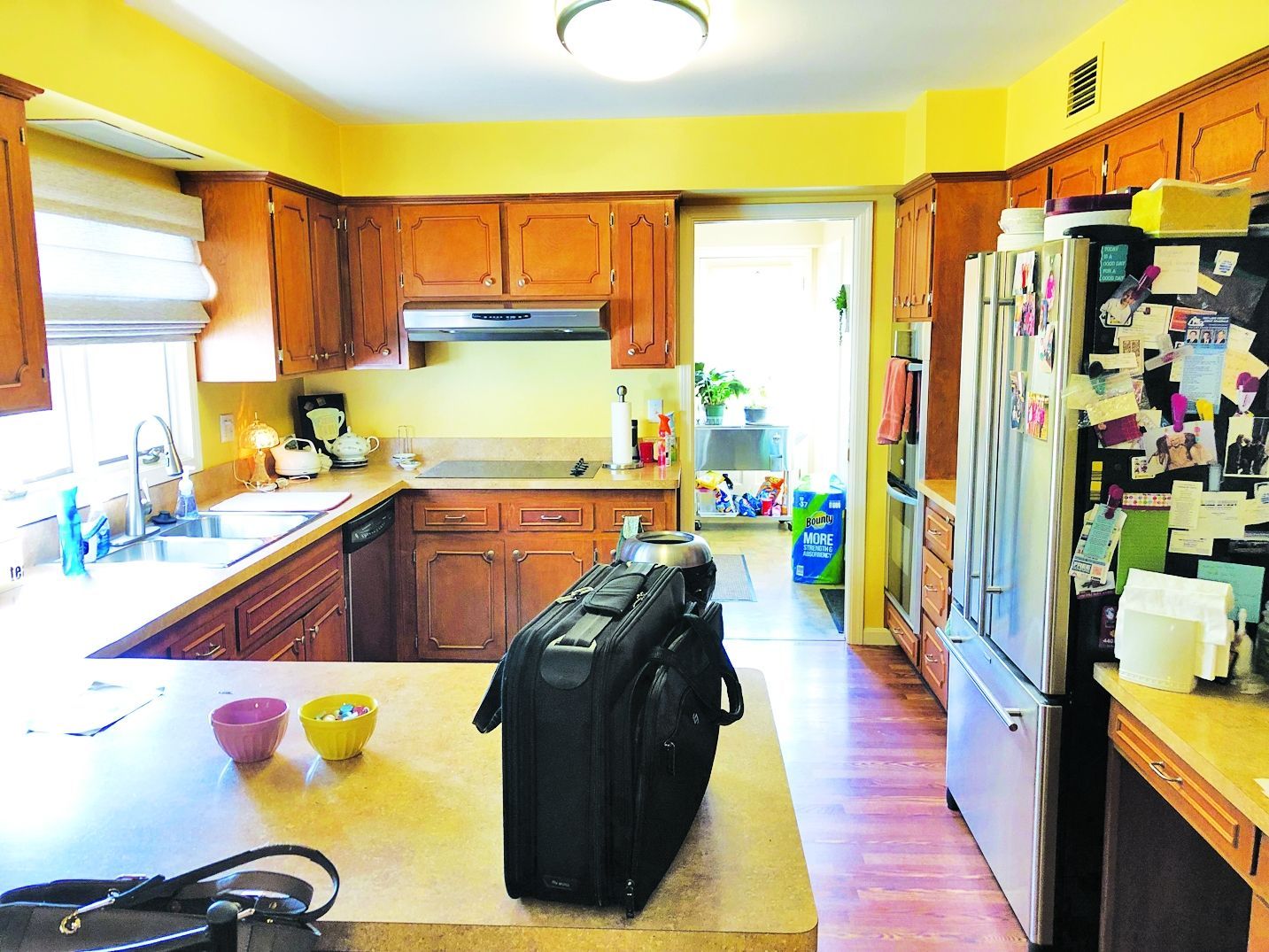 A kitchen with yellow walls, wood cabinets, a black briefcase on a counter, and a stainless steel refrigerator.