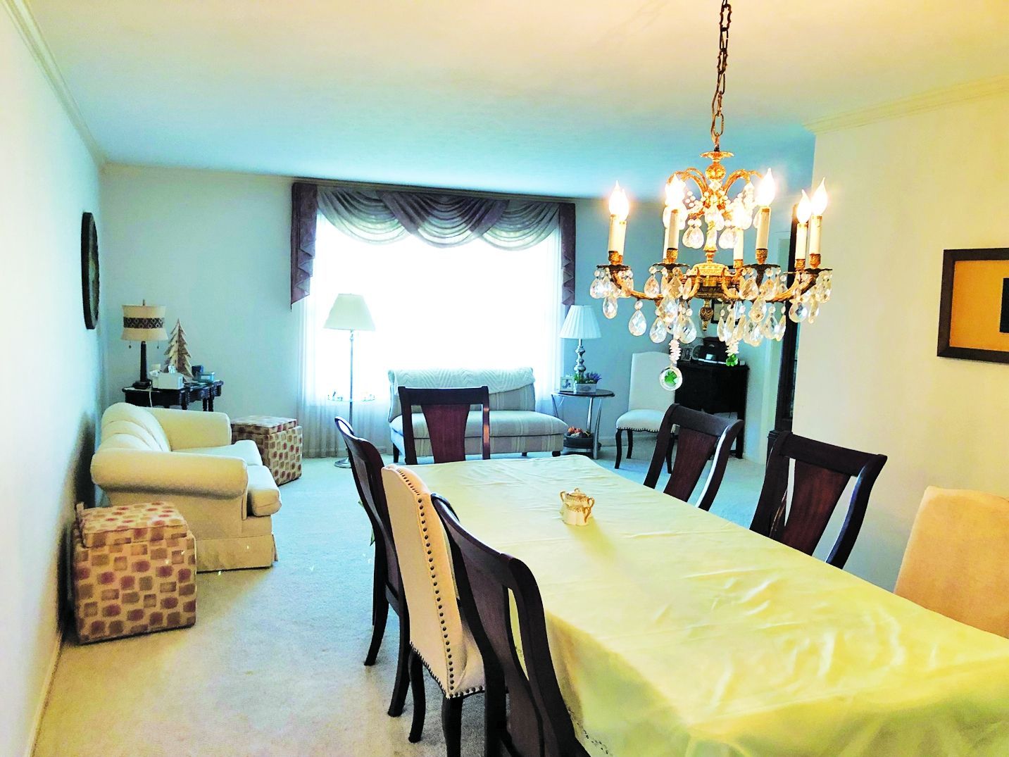 A formal dining room featuring a table with a yellow cloth and chandelier, adjacent to a living area with white furniture.