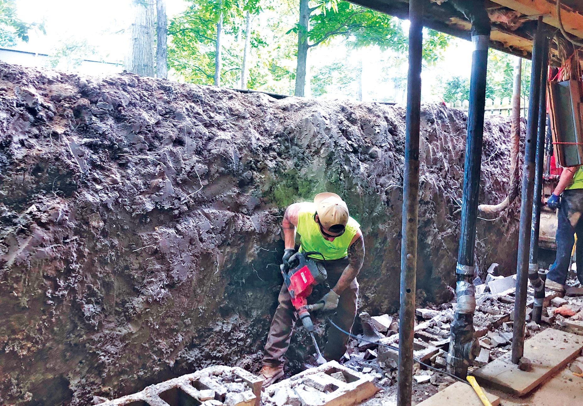 A construction worker in a high-visibility vest uses a jackhammer to break concrete in a trench supported by metal posts.
