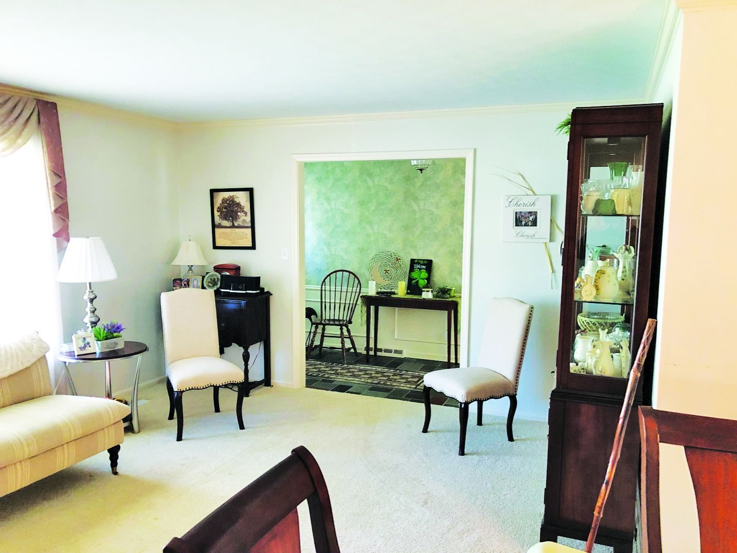 A formal living room with cream furniture, two chairs, a dark wood display cabinet, and a view into a green dining area.