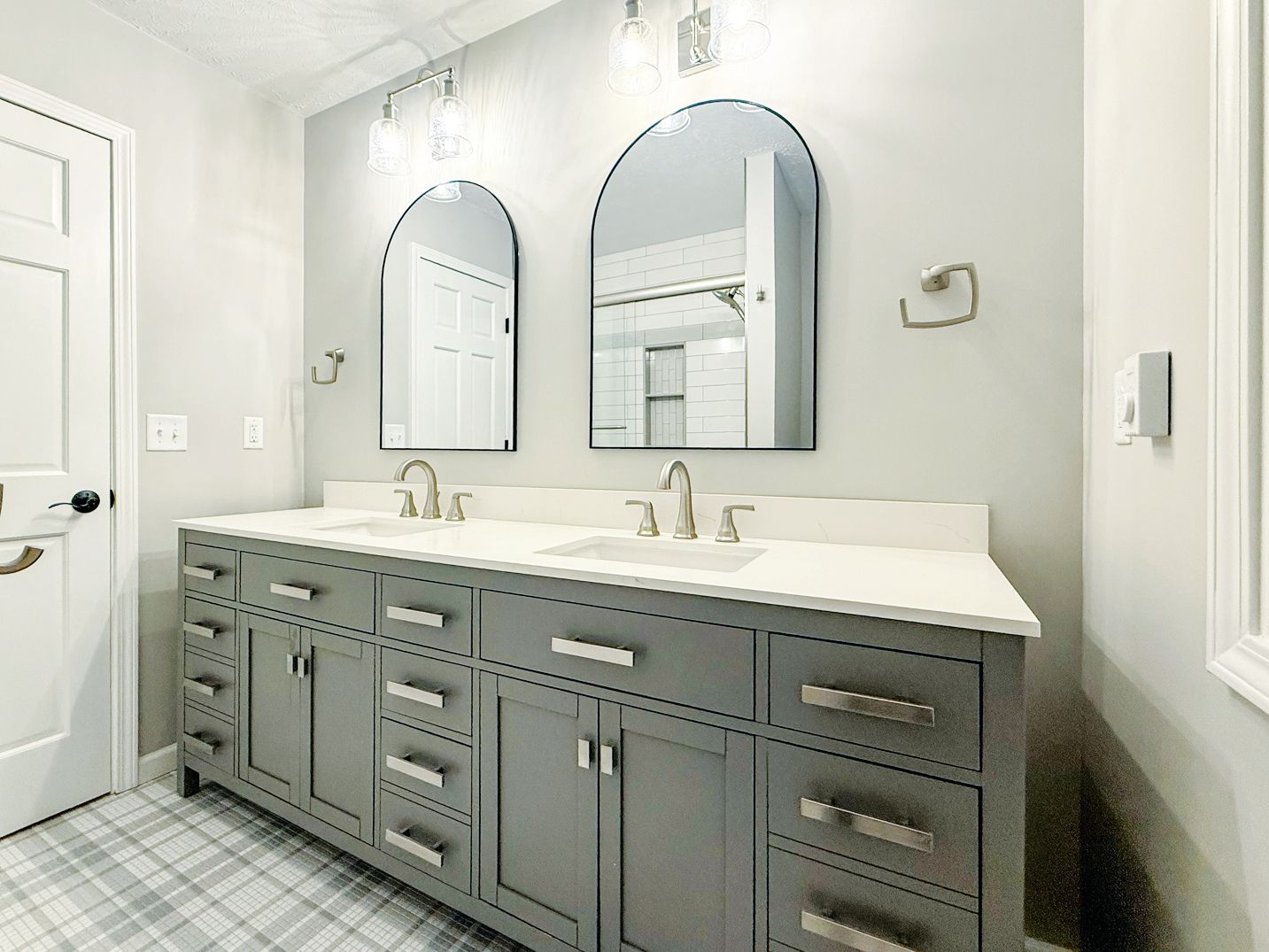 A modern bathroom featuring a gray double vanity, white countertop, dual arched mirrors, and plaid tile flooring.