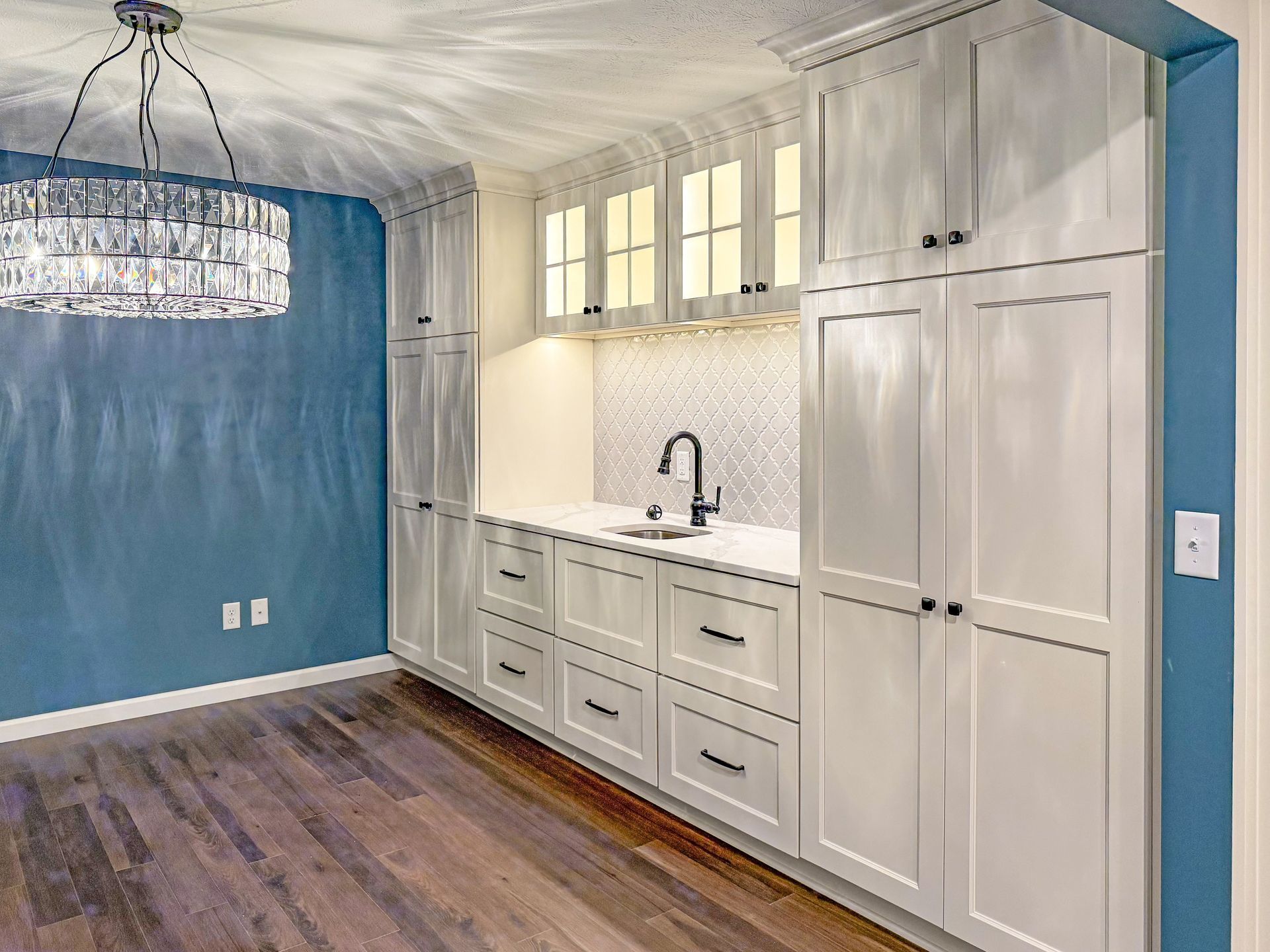 A wet bar with white cabinetry, glass-front upper cabinets, a tiled backsplash, and a large chandelier against a blue wall.