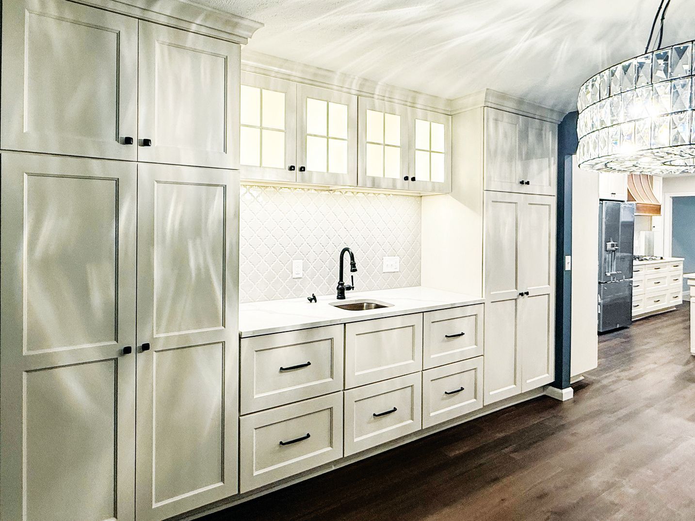 A brightly lit kitchen wet bar with white Shaker-style cabinets, a marble backsplash, black faucet, and wood flooring.
