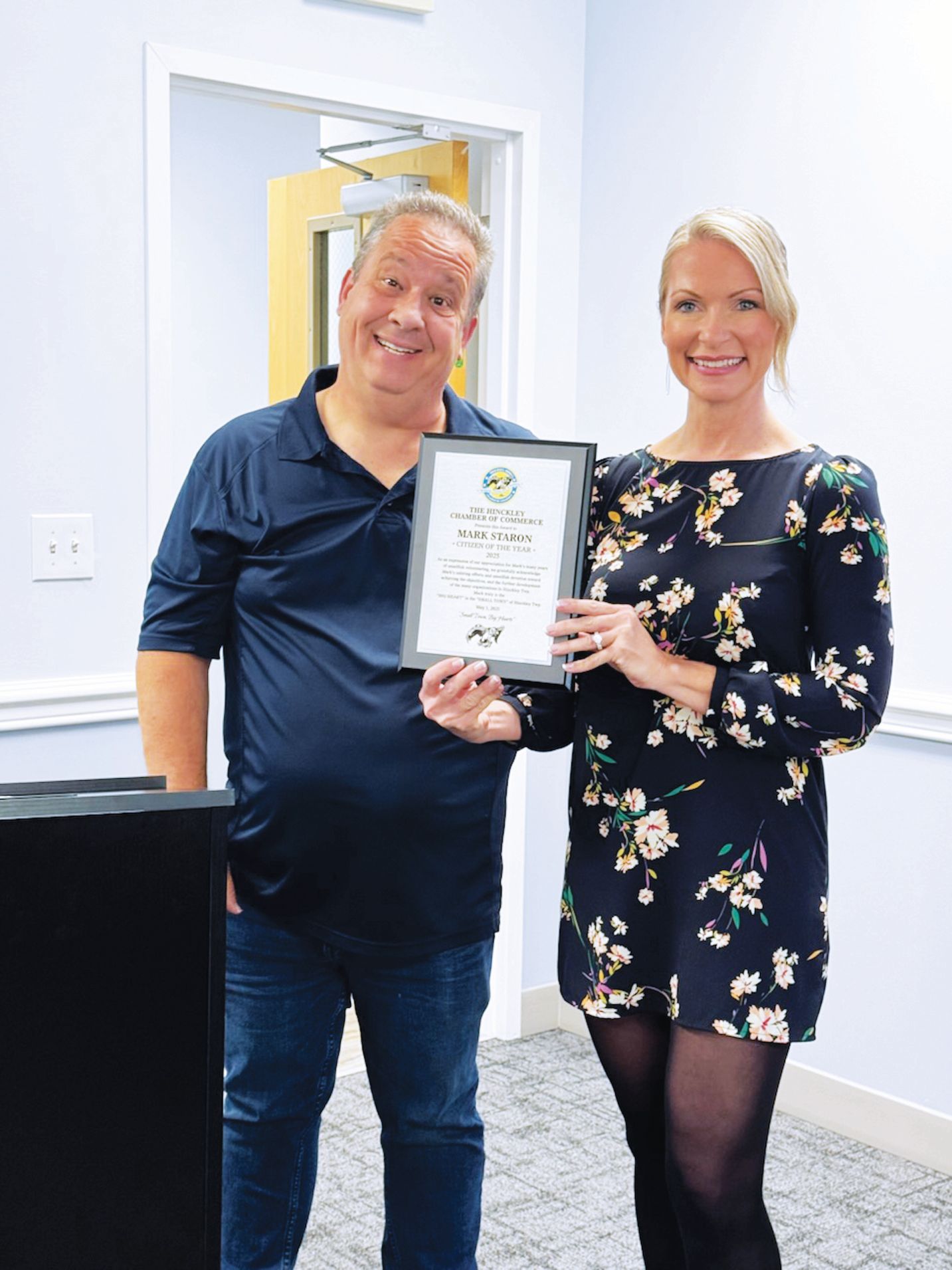 Two people smiling and holding a framed certificate in an indoor office setting.