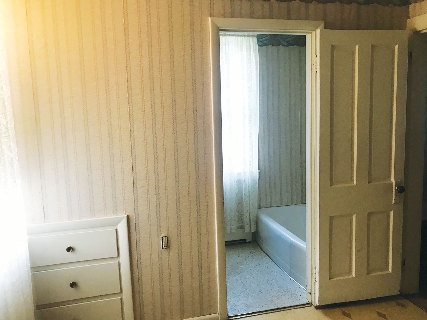 A room with striped wallpaper and a small white three-drawer unit, looking into a bathroom with a tub and a window.