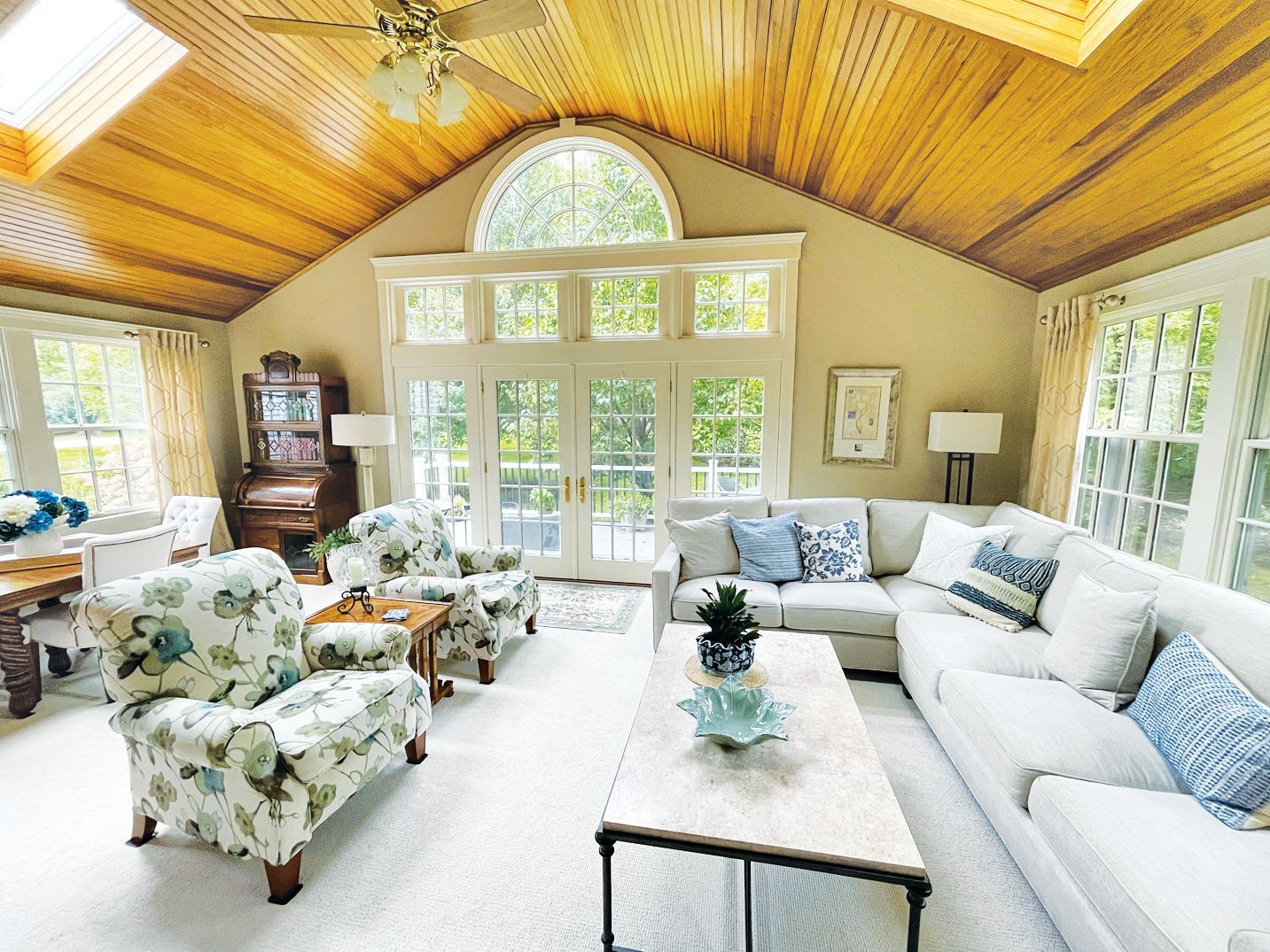 A bright, spacious sunroom features a floral armchair, white sectional sofa, vaulted wood ceiling, and large windows.