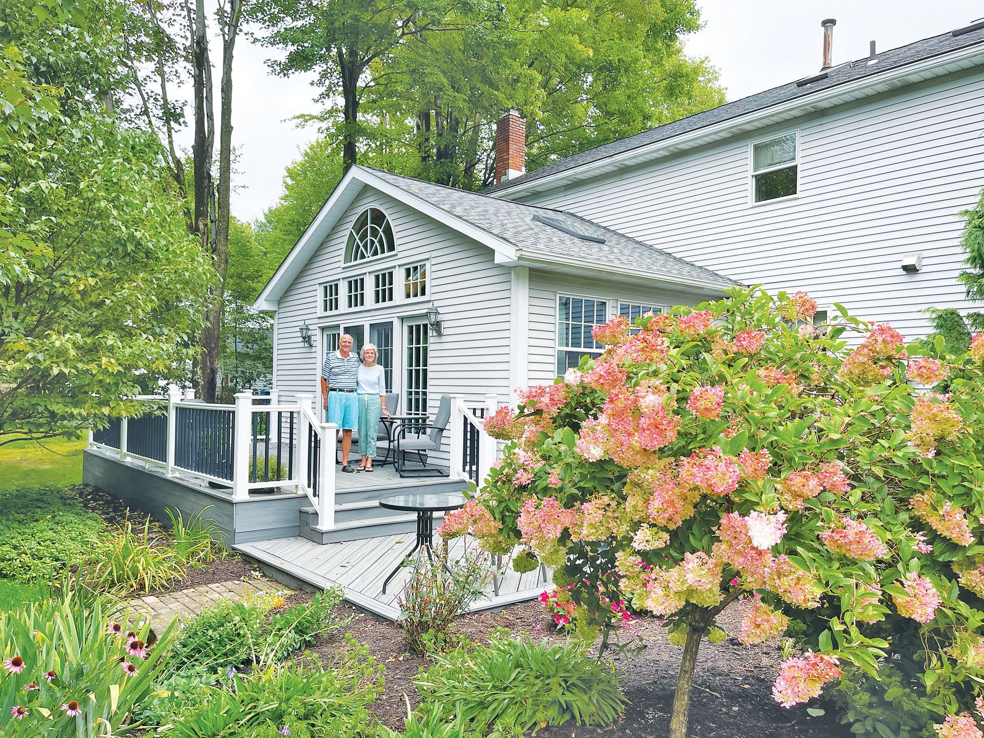An older couple stands on a gray deck outside a light gray house near a large, blooming hydrangea bush.