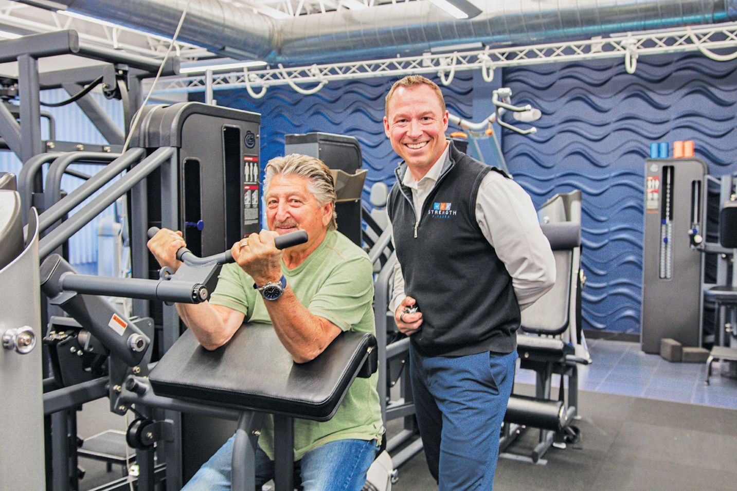A person in a light green shirt uses a weight machine, while another in a black vest stands beside them in a gym.