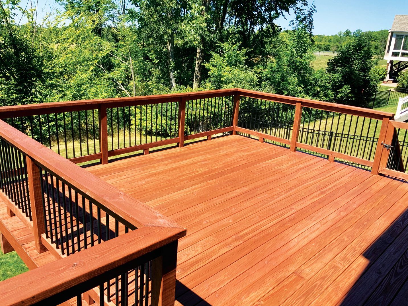 A wooden deck with a stained finish and black metal railings overlooking a lush green yard under a bright blue sky.