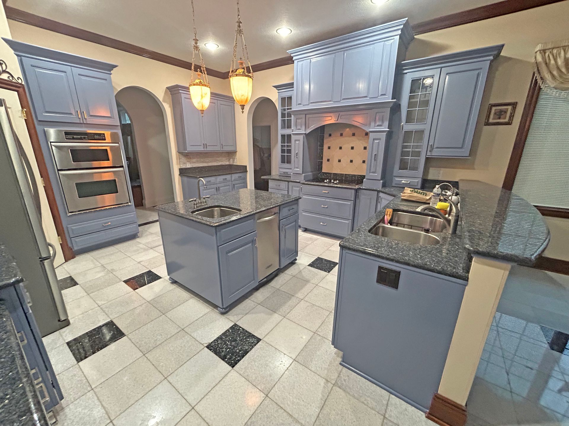 A kitchen with blue cabinets, a central island, a large range hood, stainless steel appliances, and checkered tile floor.