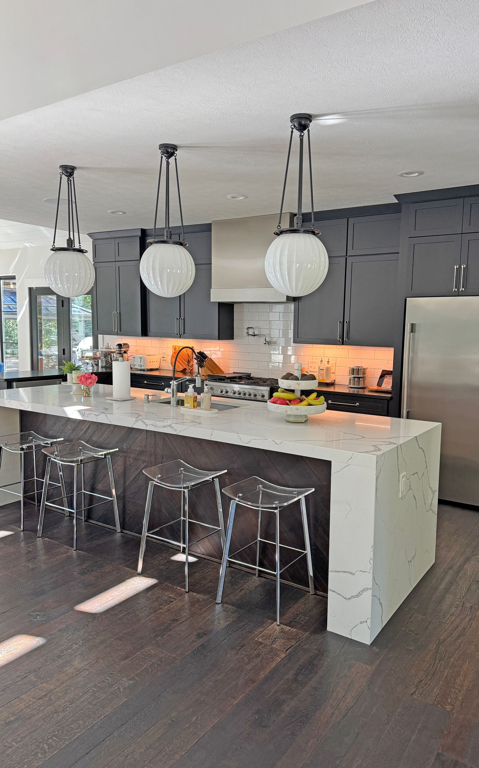 A modern kitchen with dark gray cabinets, a large white quartz island, three clear stools, and three globe pendant lights.