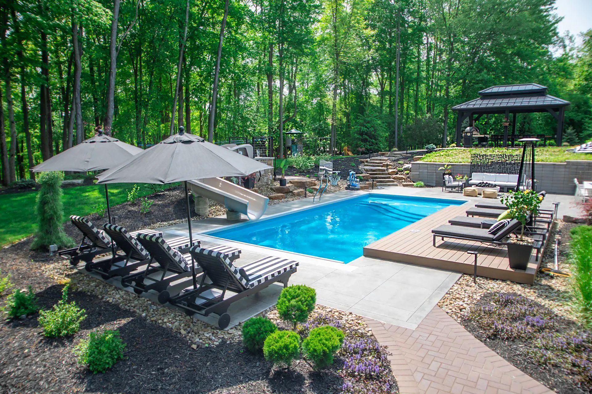An outdoor backyard with a rectangular blue swimming pool, lounge chairs under umbrellas, and a gazebo near forest trees.