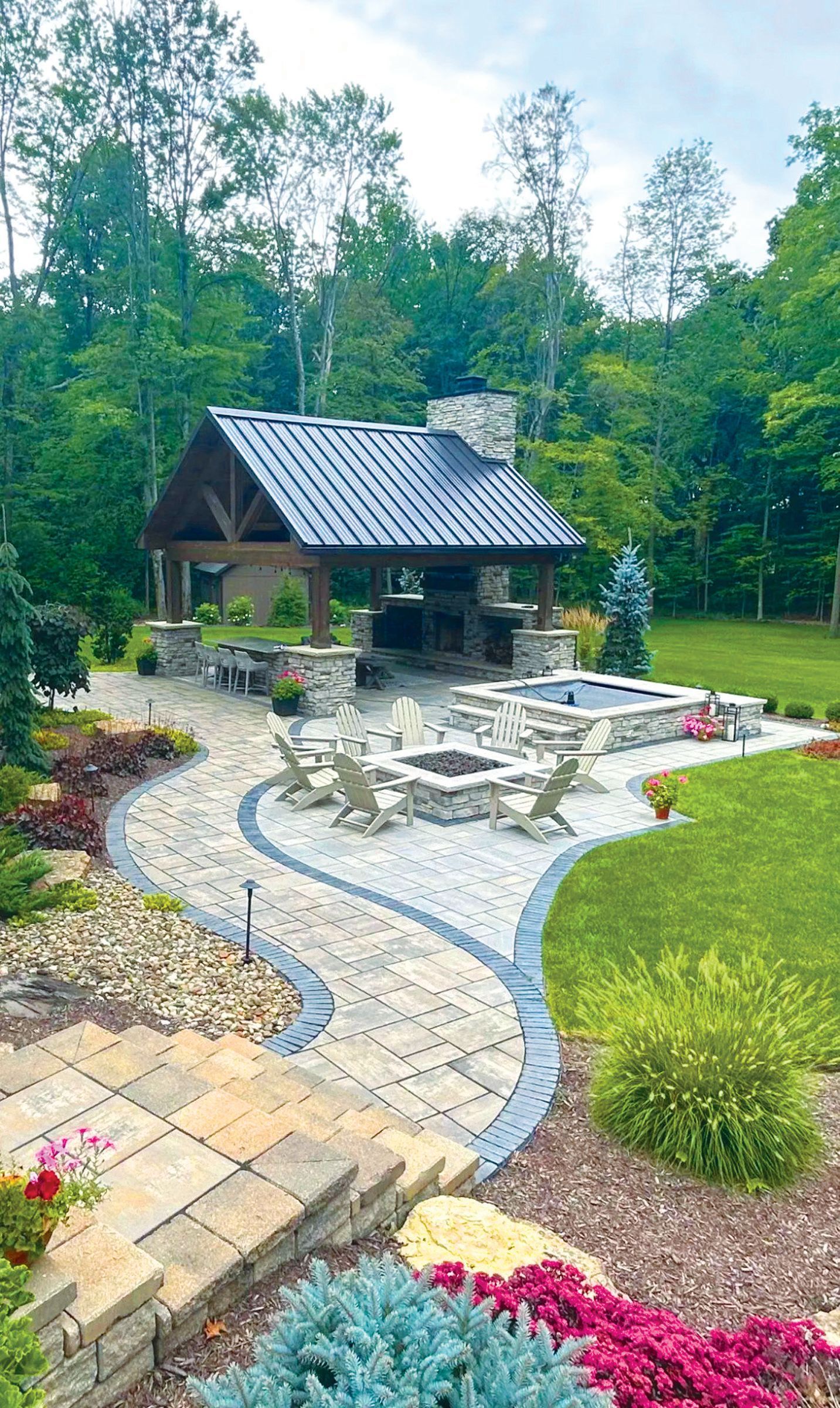 A stone patio features a fire pit, several chairs, and a covered pavilion with a fireplace, set in a lush green backyard.