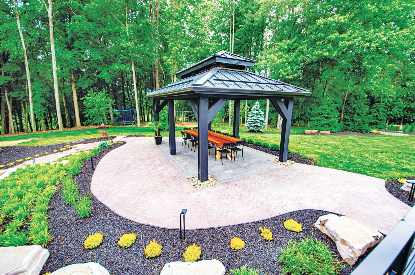A black gazebo with a dining table sits on a paved patio in a lush, green yard surrounded by trees.
