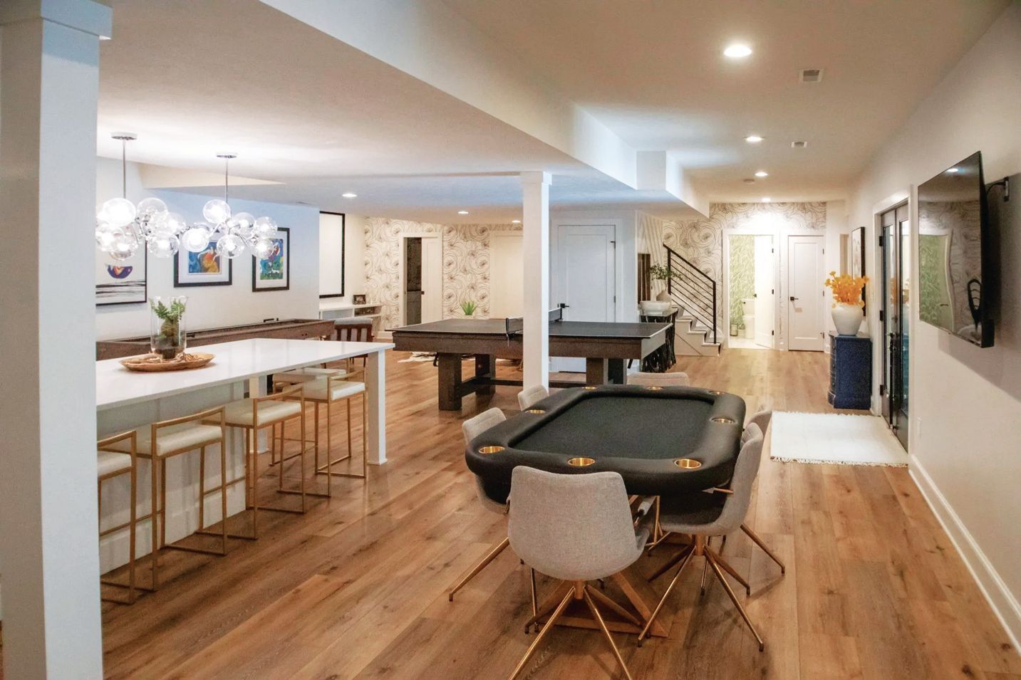 A basement recreation room with a white kitchen island, a poker table, a pool table, and light wood flooring.