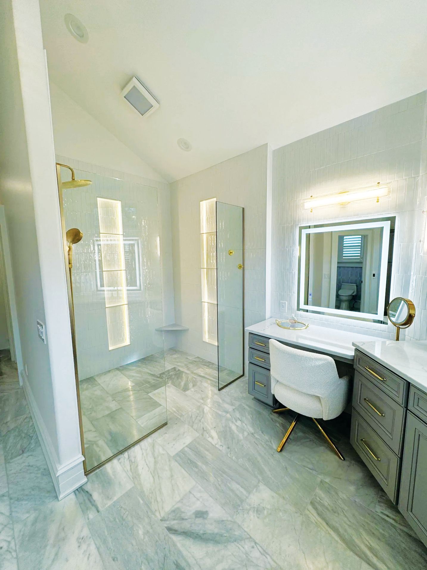 A modern marble-floored bathroom with a glass-enclosed shower, a vanity with a lighted mirror, and a white swivel chair.
