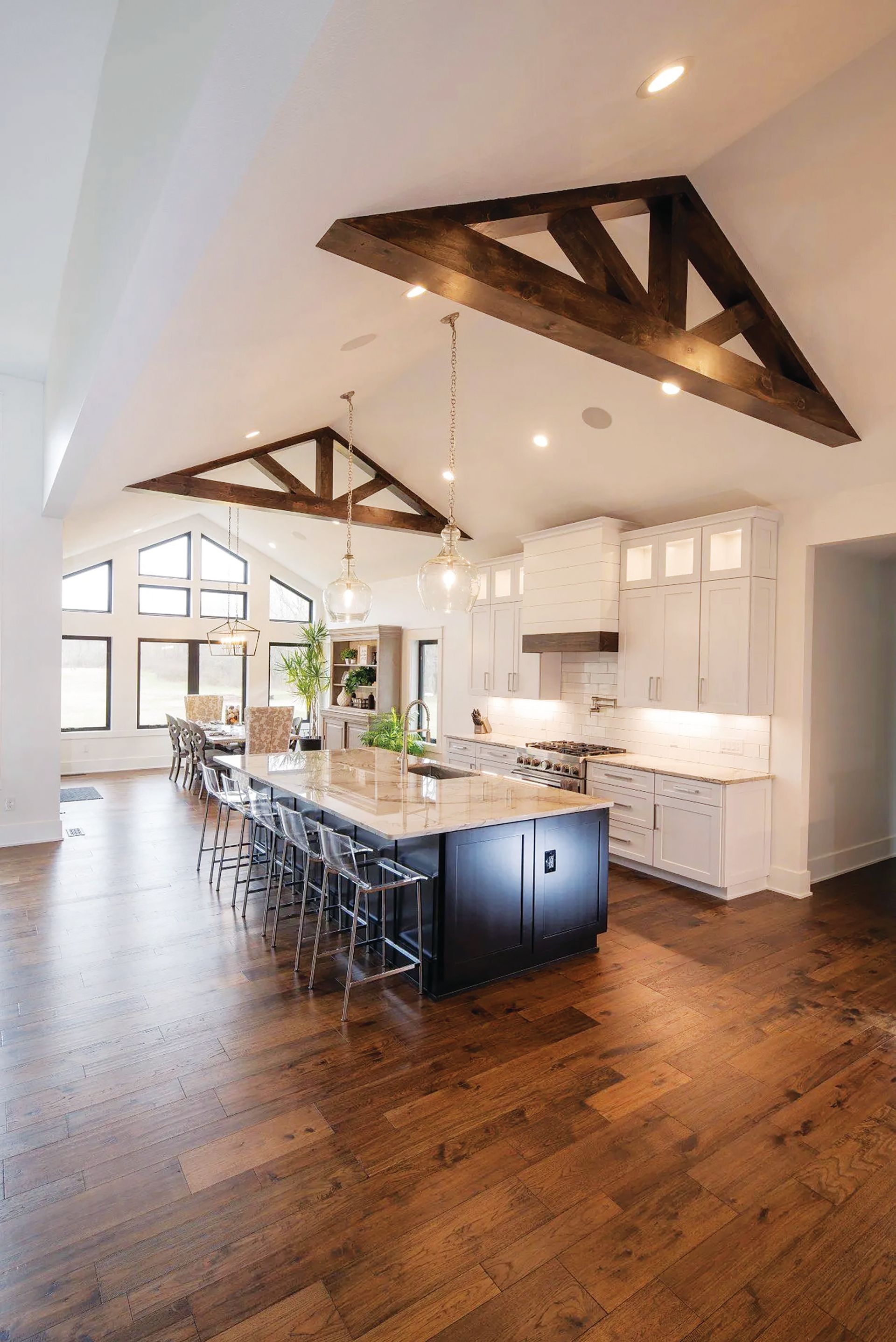 A spacious kitchen with a dark central island, white cabinetry, hardwood floors, and exposed wooden trusses on the ceiling.