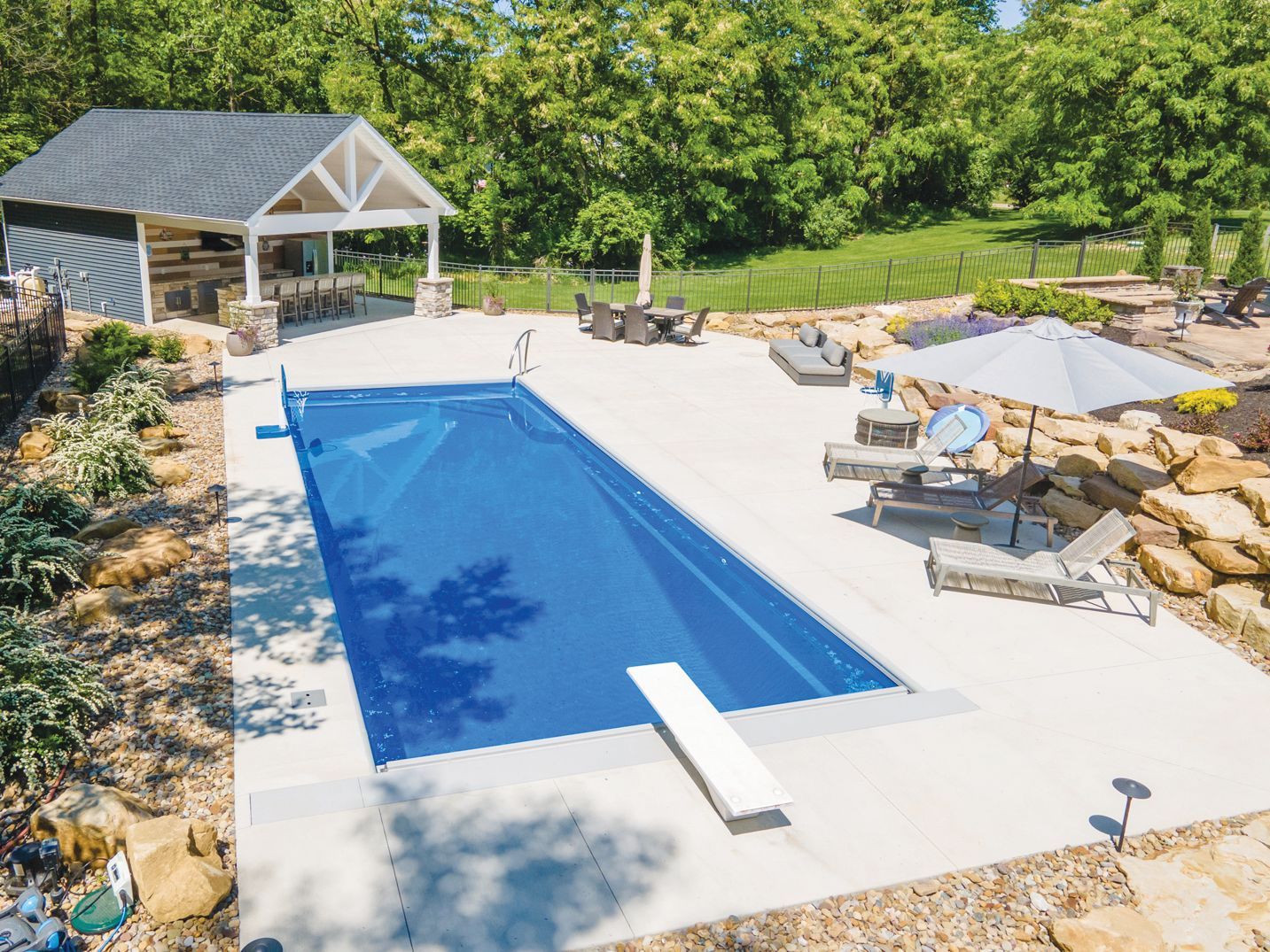 A rectangular blue swimming pool with a diving board in a sunny backyard, featuring a covered patio and lounge seating.
