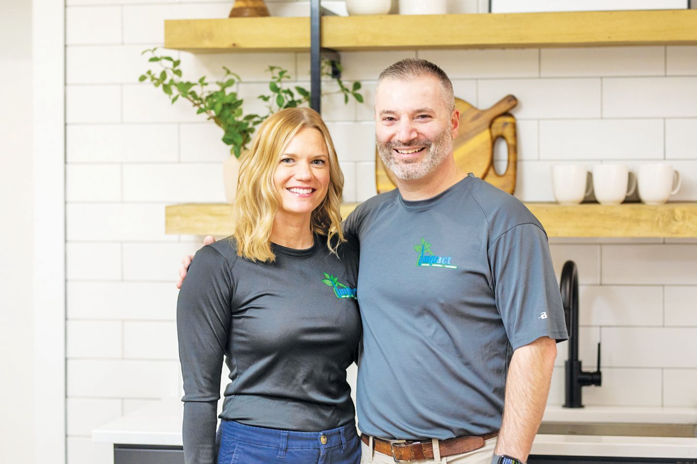 A smiling couple wearing matching grey shirts with a company logo, standing together in a kitchen with tiled walls.